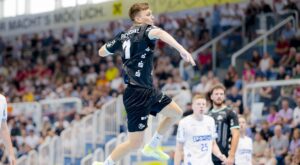 Ein Handballspieler in einem schwarzen Trikot springt in die Luft und bereitet sich auf den Wurf vor, während die Zuschauer von der Tribüne aus zusehen und die gegnerischen Spieler in weißen Trikots zuschauen.
