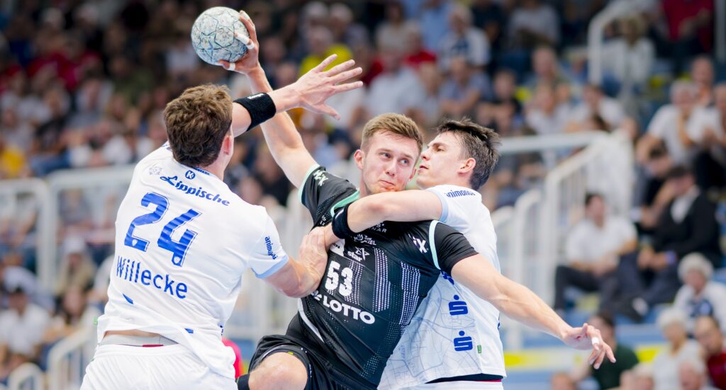 Drei männliche Handballspieler treten während eines Spiels aggressiv gegeneinander an, wobei ein Spieler in Schwarz versucht zu schießen, während zwei Spieler in Weiß eng verteidigen. Im unscharfen Hintergrund sehen Zuschauer zu.