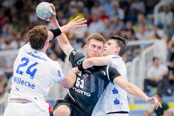 Drei männliche Handballspieler treten während eines Spiels aggressiv gegeneinander an, wobei ein Spieler in Schwarz versucht zu schießen, während zwei Spieler in Weiß eng verteidigen. Im unscharfen Hintergrund sehen Zuschauer zu.