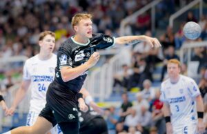 Ein Handballspieler in einem schwarzen Trikot wirft den Ball mitten im Spiel, während drei gegnerische Spieler in weißen Trikots ihn beobachten. Die Szene spielt sich in einer überfüllten Halle ab.