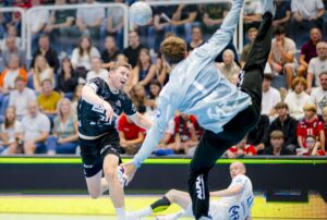 Ein Handballspieler in Schwarz springt, um den Ball zu schießen, während ein Torwart in Grau springt, um den Ball zu blocken, während im Hintergrund eine Menge von Zuschauern aufmerksam zusieht.
