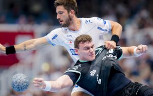 Zwei männliche Handballspieler in Aktion; einer im weißen Trikot verteidigt, während der andere im schwarzen Trikot einen Schussversuch unternimmt, wobei er den Ball hält und sich entschlossen nach vorne lehnt. Im Hintergrund ist eine unscharfe Menschenmenge zu sehen.