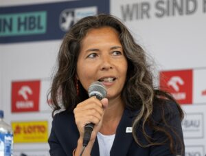 Eine Frau mit langen, gewellten braunen und grauen Haaren spricht bei einer Pressekonferenz in ein Mikrofon. Sie trägt einen marineblauen Blazer und sitzt vor einer Kulisse mit verschiedenen Logos und Texten.