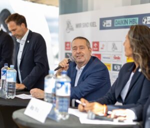 Ein Mann in einem blauen Anzug spricht an einem Pressekonferenztisch in ein Mikrofon. Neben ihm sitzen zwei weitere Personen, hinter denen Sponsorenlogos auf einem Hintergrund zu sehen sind. Flaschen mit Wasser stehen auf dem Tisch.