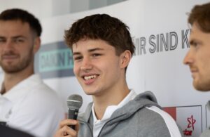Ein junger Mann in einem grauen Kapuzenpulli lächelt, während er bei einer Veranstaltung in ein Mikrofon spricht. Neben ihm sind zwei weitere Männer vor einem Hintergrund mit Texten und Logos zu sehen.