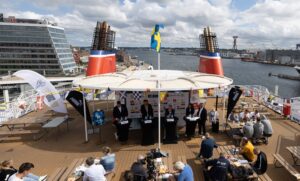 Auf dem Deck einer großen Fähre findet ein Schachturnier statt. Auf der Bühne sind vier Schachbretter mit Spielern und Schiedsrichtern aufgebaut, während die Zuschauer drum herum sitzen. Darüber weht die schwedische Flagge, und im Hintergrund ist der Hafen mit Gebäuden zu sehen.