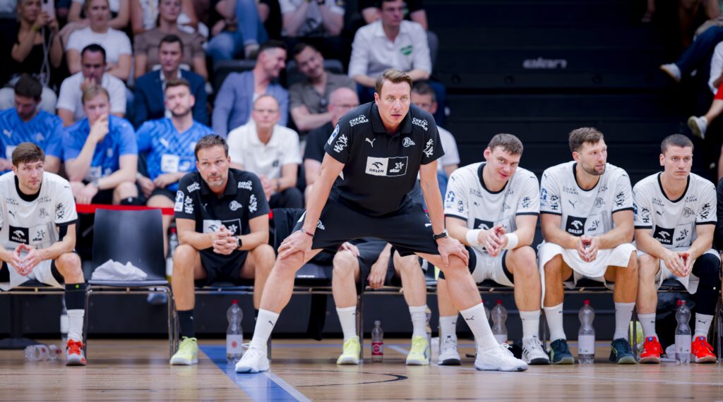 Ein Handballtrainer in schwarzer Sportkleidung steht vor einer Bank und beobachtet aufmerksam das Spiel, während fünf Spieler in weißer Kleidung neben ihm sitzen. Die Zuschauer füllen die Tribünen im Hintergrund.