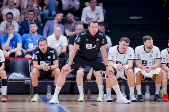 Ein Handballtrainer in schwarzer Sportkleidung steht vor einer Bank und beobachtet aufmerksam das Spiel, während fünf Spieler in weißer Kleidung neben ihm sitzen. Die Zuschauer füllen die Tribünen im Hintergrund.