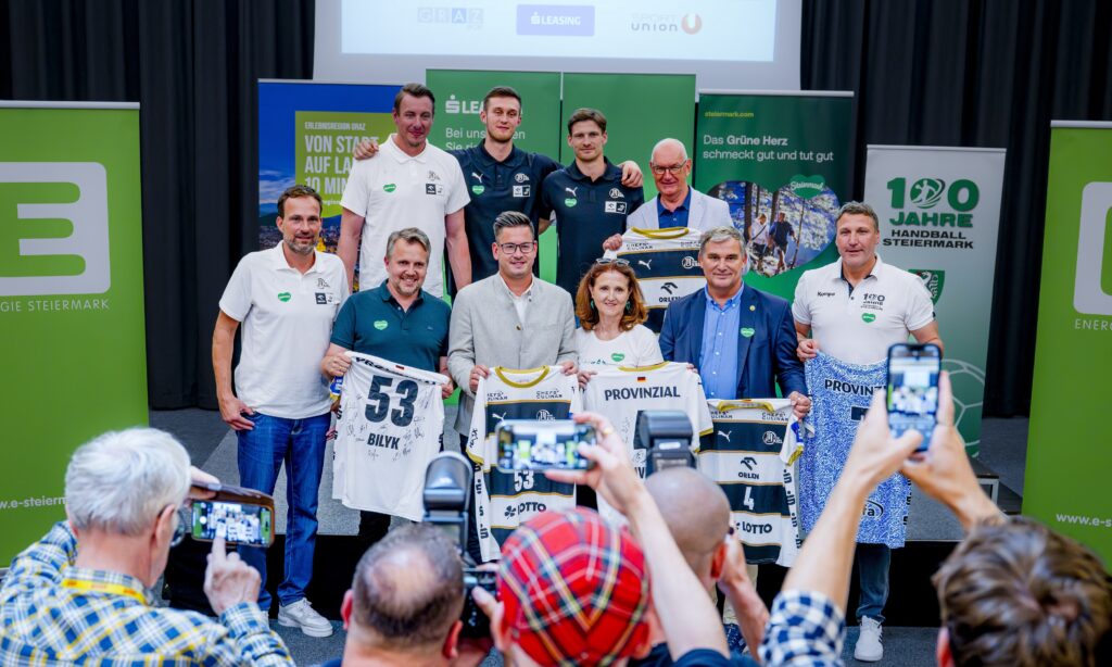 Eine Gruppe von Menschen posiert drinnen bei einer Veranstaltung und hält Sporttrikots mit der Nummer 53 hoch. Einige Teilnehmer machen Fotos, während andere vor grünen Bannern und einem großen Bildschirm stehen und lächeln.