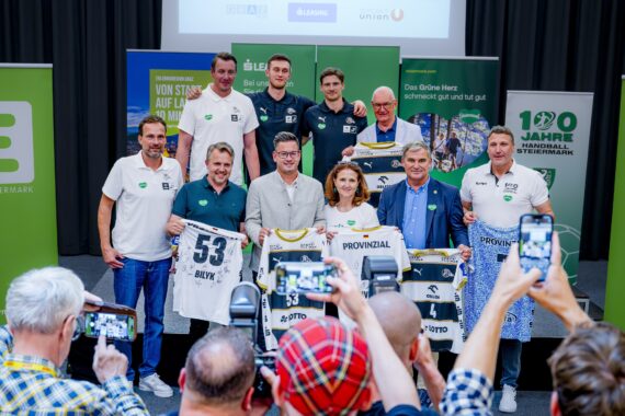 Eine Gruppe von Menschen posiert drinnen bei einer Veranstaltung und hält Sporttrikots mit der Nummer 53 hoch. Einige Teilnehmer machen Fotos, während andere vor grünen Bannern und einem großen Bildschirm stehen und lächeln.