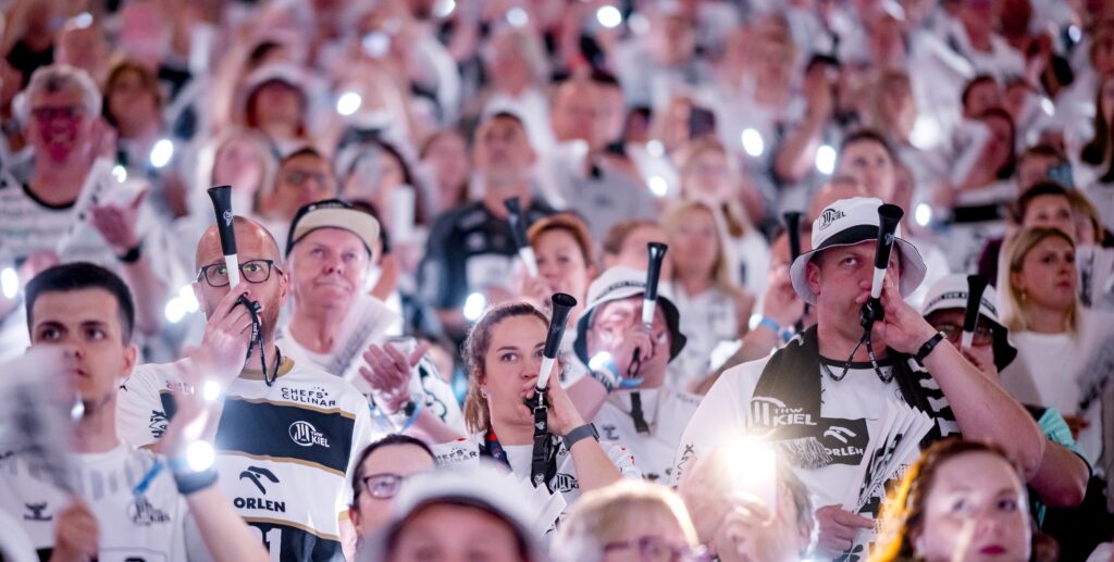 HANDBALL 2024/25: Maschinensucher EHF Finals Eine große Menge begeisterter, weiß gekleideter Fans, die leuchtende Lichter in die Höhe halten und in Tröten blasen, füllt die Tribünen bei einer Sportveranstaltung.