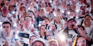 Eine große Menge begeisterter, weiß gekleideter Fans, die leuchtende Lichter in die Höhe halten und in Tröten blasen, füllt die Tribünen bei einer Sportveranstaltung.