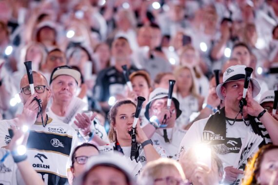 Eine große Menge begeisterter, weiß gekleideter Fans, die leuchtende Lichter in die Höhe halten und in Tröten blasen, füllt die Tribünen bei einer Sportveranstaltung.