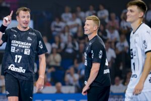 Drei männliche Handballspieler in schwarz-weißen Trikots stehen während eines Spiels auf dem Spielfeld. Ein Spieler gestikuliert mit erhobenem Finger, während die anderen lächelnd zusehen. Das Publikum ist im unscharfen Hintergrund zu sehen.