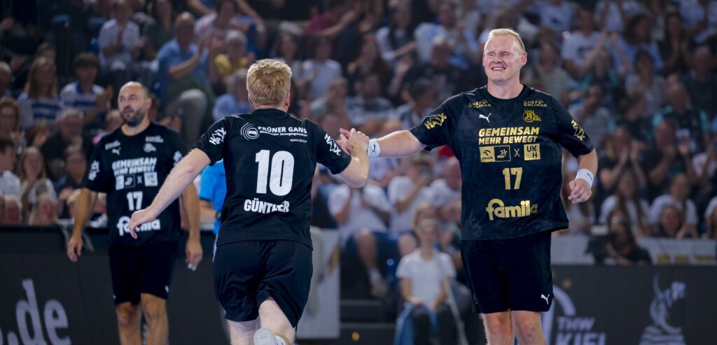 Zwei Handballspieler in schwarzen Trikots klatschen sich während eines Spiels gegenseitig ab, während im Hintergrund eine jubelnde Menge steht. Auf dem Trikot des einen Spielers steht die Nummer 10, auf dem des anderen die 17.