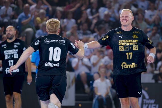 Zwei Handballspieler in schwarzen Trikots klatschen sich während eines Spiels gegenseitig ab, während im Hintergrund eine jubelnde Menge steht. Auf dem Trikot des einen Spielers steht die Nummer 10, auf dem des anderen die 17.