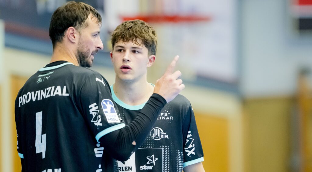 Zwei männliche Handballspieler in schwarzen Trikots, einer mit der Nummer 4, stehen in einer Halle und führen ein ernstes Gespräch. Der ältere Spieler gestikuliert mit seinem Finger, während der jüngere Spieler aufmerksam zuhört.