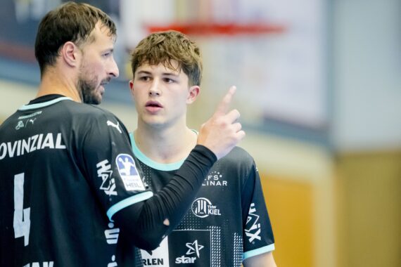 Zwei männliche Handballspieler in schwarzen Trikots, einer mit der Nummer 4, stehen in einer Halle und führen ein ernstes Gespräch. Der ältere Spieler gestikuliert mit seinem Finger, während der jüngere Spieler aufmerksam zuhört.