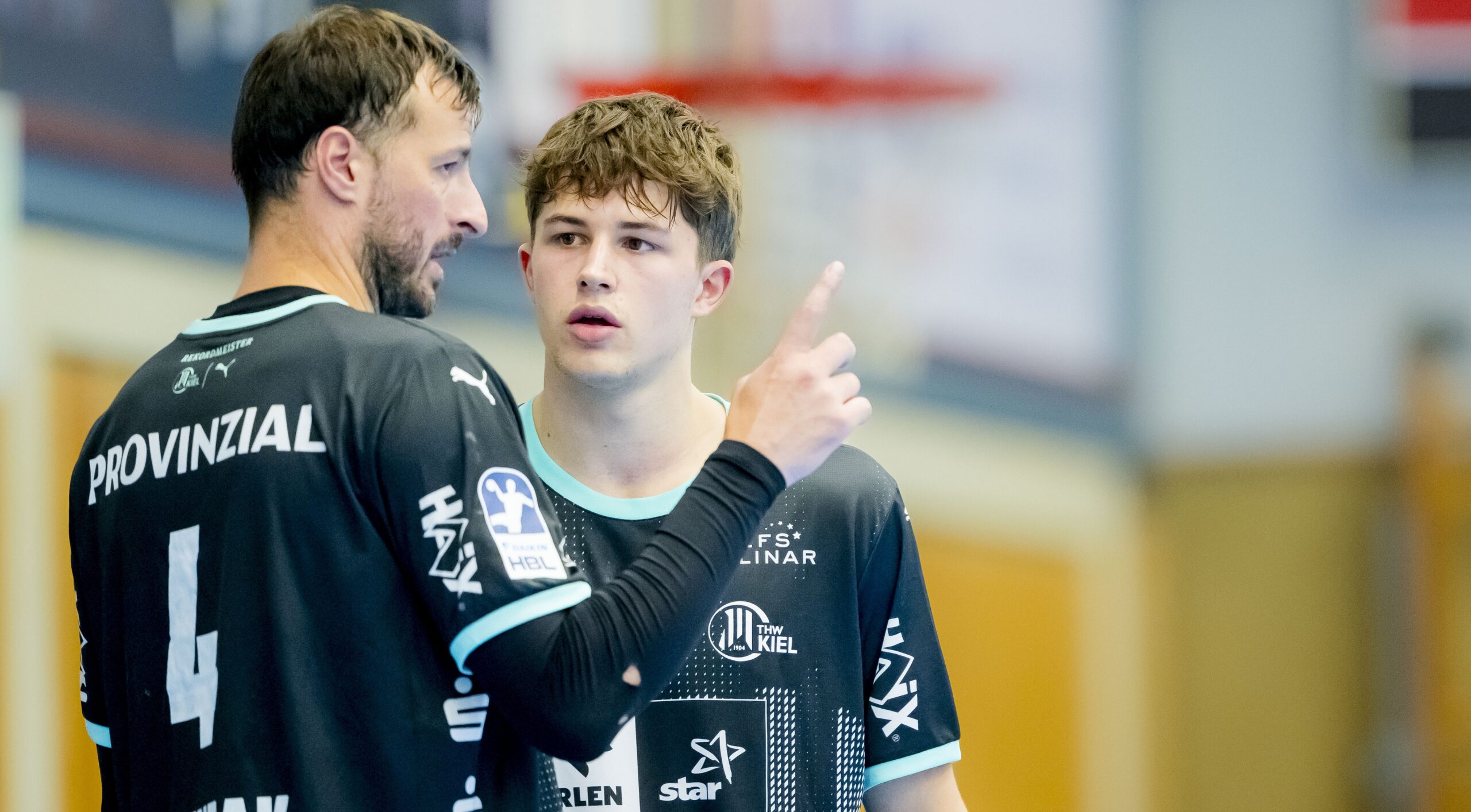 Zwei männliche Handballspieler in schwarzen Trikots, einer mit der Nummer 4, stehen in einer Halle und führen ein ernstes Gespräch. Der ältere Spieler gestikuliert mit seinem Finger, während der jüngere Spieler aufmerksam zuhört.