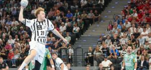 Ein Handballspieler in einem weiß-schwarzen Trikot springt in der Luft, um den Ball zu werfen, während andere Spieler und ein großes Publikum im Hintergrund in einer Halle zusehen.