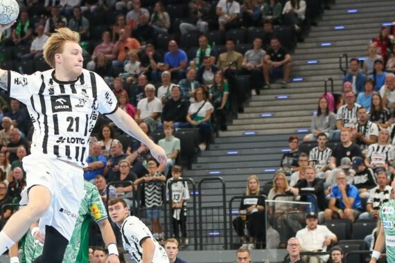 Ein Handballspieler in einem weiß-schwarzen Trikot springt in der Luft, um den Ball zu werfen, während andere Spieler und ein großes Publikum im Hintergrund in einer Halle zusehen.