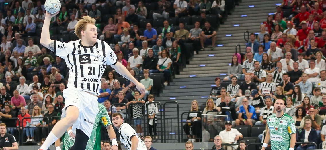 Ein Handballspieler in einem weiß-schwarzen Trikot springt in der Luft, um den Ball zu werfen, während andere Spieler und ein großes Publikum im Hintergrund in einer Halle zusehen.