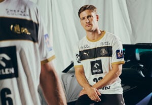 Ein männlicher Fußballspieler in einem weiß-goldenen Trikot sitzt auf einem Auto und blickt nach vorne. Ein anderer Spieler, der nur teilweise sichtbar und unscharf ist, steht mit dem Rücken zugewandt im Vordergrund. Der Hintergrund besteht aus drapiertem Stoff.