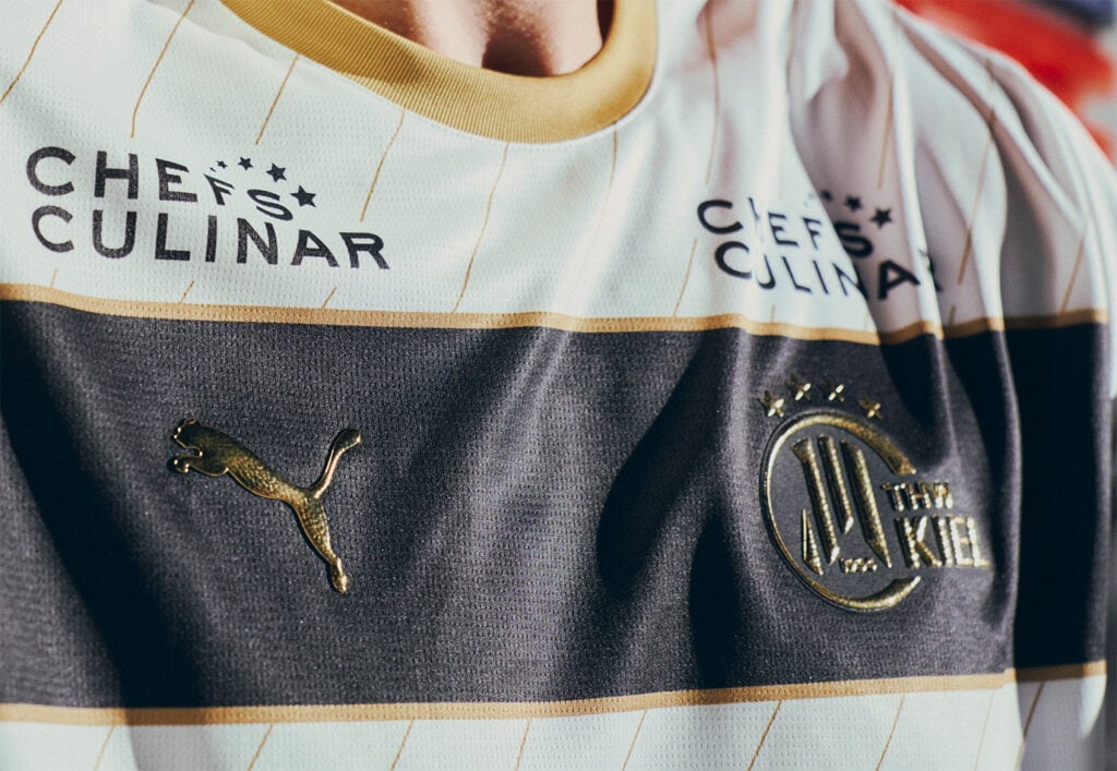 Nahaufnahme eines Fußballtrikots mit goldenen Akzenten, dem Puma-Logo, dem Vereinswappen von Holstein Kiel und dem Sponsorenlogo von CHEFS CULINAR auf weißem und dunklem, horizontal gestreiftem Hintergrund.