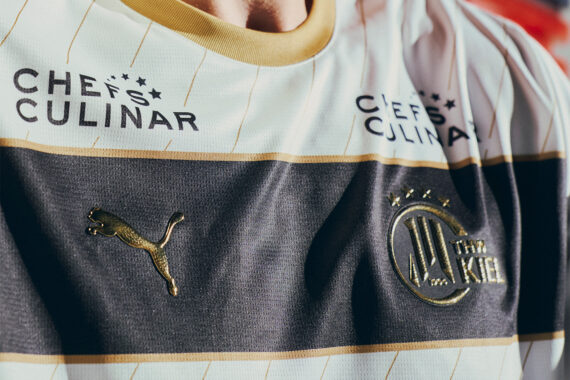 Nahaufnahme eines Fußballtrikots mit goldenen Akzenten, dem Puma-Logo, dem Vereinswappen von Holstein Kiel und dem Sponsorenlogo von CHEFS CULINAR auf weißem und dunklem, horizontal gestreiftem Hintergrund.