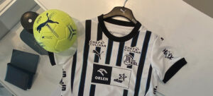 Ein weiß-schwarz gestreiftes Fußballtrikot auf einem Bügel neben einem neongelben Puma-Fußball auf einem Schreibtisch. Das Trikot zeigt mehrere Sponsorenlogos und den Namen THW Kiel auf der Brust.