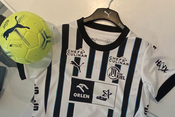 Ein weiß-schwarz gestreiftes Fußballtrikot auf einem Bügel neben einem neongelben Puma-Fußball auf einem Schreibtisch. Das Trikot zeigt mehrere Sponsorenlogos und den Namen THW Kiel auf der Brust.