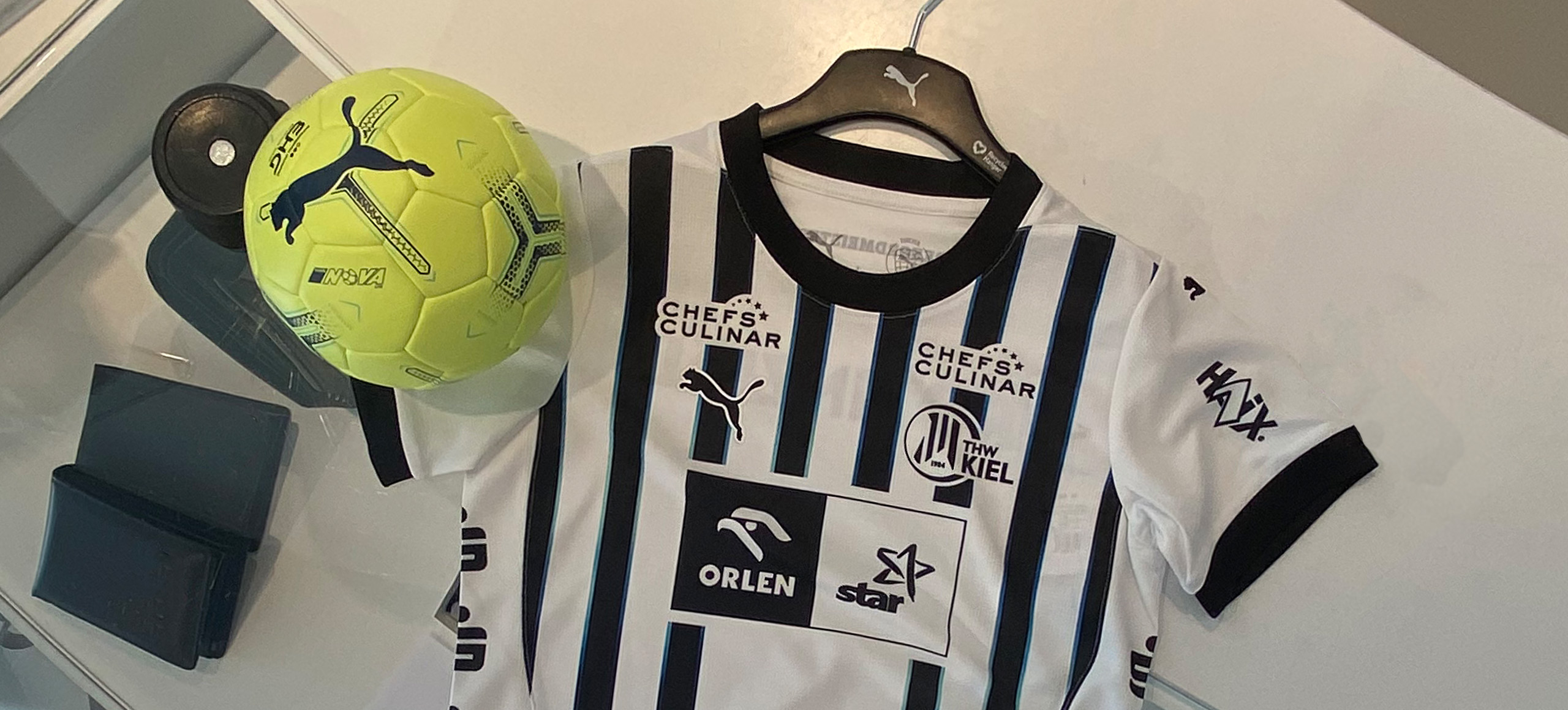 Ein weiß-schwarz gestreiftes Fußballtrikot auf einem Bügel neben einem neongelben Puma-Fußball auf einem Schreibtisch. Das Trikot zeigt mehrere Sponsorenlogos und den Namen THW Kiel auf der Brust.