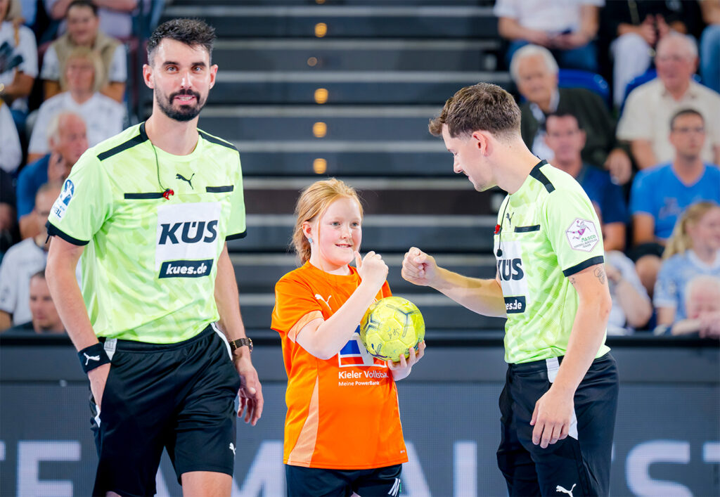 Ein junges Mädchen in einem orangefarbenen T-Shirt, das auf eine Bewerbung als THW Kiel Ballkind hofft, hält einen Handball und stößt einen Schiedsrichter mit der Faust an, während ein anderer Schiedsrichter in Grün in der Nähe steht. Im Hintergrund sind Zuschauer zu sehen.