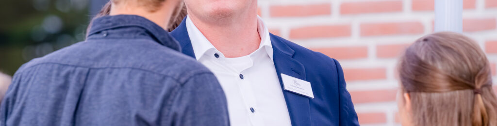 Drei Personen unterhalten sich, wobei nur die Unterseiten der Gesichter und Oberkörper zu sehen sind. Eine Person trägt eine Anzugsjacke mit einem Namensschild, die anderen sind leger gekleidet. Im Hintergrund ist eine Backsteinmauer zu sehen.