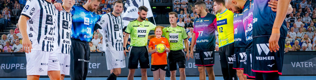 Eine Gruppe von Handballspielern in schwarz-weißen und blauen Trikots steht zusammen mit Schiedsrichtern und einem THW-Bewerbungs-Ballkind, das einen Ball hält, auf einem Hallenplatz vor einem Spiel, mit einer großen Menschenmenge im Hintergrund.