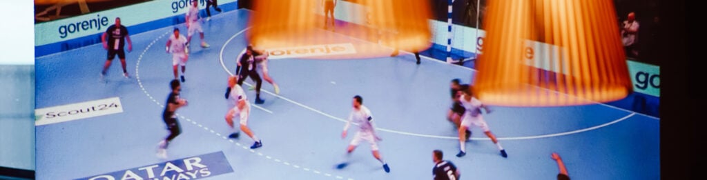 2526_Titel_Partner-Viewing Ein Handballspiel auf einem blauen Spielfeld, mit Spielern in weißen und dunklen Trikots; zwei orangefarbene Scheinwerfer sind über das Bild gelegt.