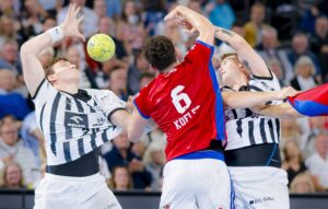 Drei männliche Handballspieler liefern sich einen intensiven Wettkampf; einer im roten Trikot (Nummer 6) versucht zu schießen, während zwei Spieler in schwarz-weißen Trikots ihn abblocken, während sich ein gelber Ball in der Luft befindet und das Publikum im Hintergrund verschwommen ist.