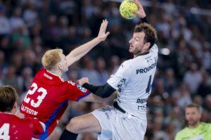 Ein Handballspieler in einem weißen Trikot springt, um den Ball zu schießen, während ein Verteidiger in einem roten Trikot versucht, ihn zu blockieren. Im Hintergrund sieht man eine unscharfe Zuschauermenge, die das Spiel verfolgt.