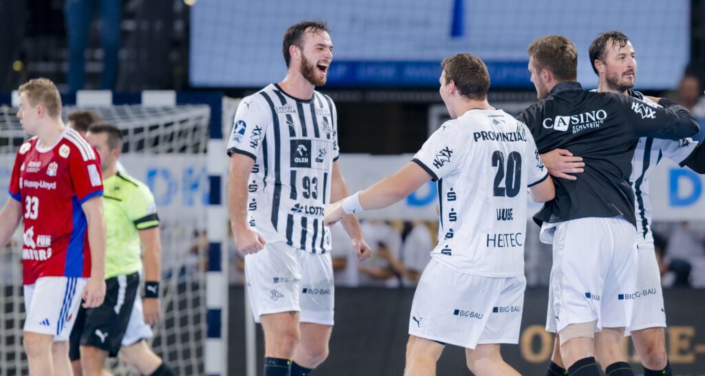 Vier Handballspieler in weißen Trikots feiern auf dem Spielfeld, lächeln und umarmen sich, während zwei Spieler in roten Trikots und ein Schiedsrichter im Hintergrund in der Nähe des Tores davonlaufen.