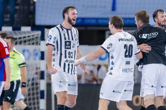 Vier Handballspieler in weißen Trikots feiern auf dem Spielfeld, lächeln und umarmen sich, während zwei Spieler in roten Trikots und ein Schiedsrichter im Hintergrund in der Nähe des Tores davonlaufen.
