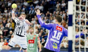Ein Handballspieler in einem schwarz-weiß gestreiften Trikot springt, um den Ball in Richtung Tor zu werfen, während ein Torwart in einem lila Trikot und ein weiterer Verteidiger in Grün sich darauf vorbereiten, ihn zu blockieren. Die Szene spielt sich in einer überfüllten Arena ab.