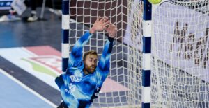 Ein männlicher Handballtorwart in einem blauen Trikot springt mit erhobenen Armen, um einen auf das Tor zukommenden Ball abzuwehren.