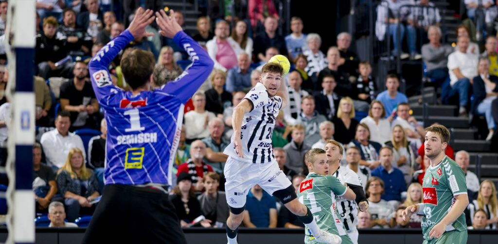 Ein Handballspieler in einem weiß-schwarzen Trikot springt, um den Ball in Richtung Tor zu werfen, während der gegnerische Torwart und die Verteidiger in Grün sich darauf vorbereiten, ihn zu blockieren. Das Publikum im Hintergrund schaut aufmerksam zu.
