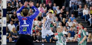 Ein Handballspieler in einem weiß-schwarzen Trikot springt, um den Ball in Richtung Tor zu werfen, während der gegnerische Torwart und die Verteidiger in Grün sich darauf vorbereiten, ihn zu blockieren. Das Publikum im Hintergrund schaut aufmerksam zu.
