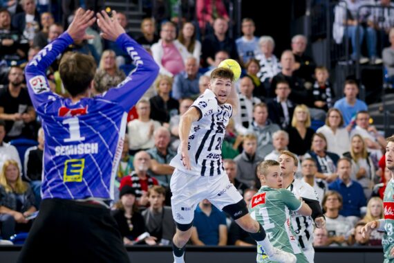 Ein Handballspieler in einem weiß-schwarzen Trikot springt, um den Ball in Richtung Tor zu werfen, während der gegnerische Torwart und die Verteidiger in Grün sich darauf vorbereiten, ihn zu blockieren. Das Publikum im Hintergrund schaut aufmerksam zu.