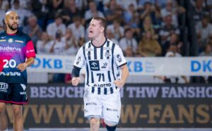 Ein Handballspieler in einem weiß-schwarz gestreiften Trikot jubelt auf dem Spielfeld, während ein anderer Spieler in einem rosa-schwarzen Trikot vorbeiläuft. Die Zuschauer füllen die Tribünen im Hintergrund.