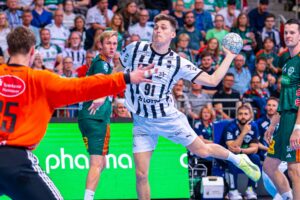 Ein männlicher Handballspieler in einem weiß-schwarz gestreiften Trikot springt, um den Ball in Richtung Tor zu werfen, während der Torwart in einem orangefarbenen Trikot und andere Spieler in grünen Trikots aufmerksam zusehen. Die Zuschauer sitzen im Hintergrund.