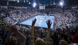 Eine große Menschenmenge in einer Halle jubelt und reißt die Hände in die Höhe, während sie ein Handballspiel auf einem Spielfeld verfolgt, das von hellen Lichtern beleuchtet wird und in dem Tausende von Zuschauern sitzen.
