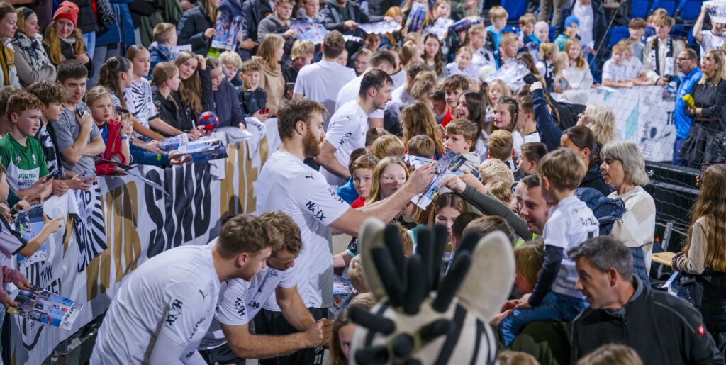 Sportler in weißen Trikots geben Autogramme und unterhalten sich mit einer großen Menge lächelnder Fans, darunter auch Kinder, bei einer Hallenveranstaltung. Die Leute halten Poster und Fanartikel in der Hand, und im Vordergrund ist ein Zebra-Maskottchen zu sehen.