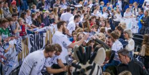 Sportler in weißen Trikots geben Autogramme und unterhalten sich mit einer großen Menge lächelnder Fans, darunter auch Kinder, bei einer Hallenveranstaltung. Die Leute halten Poster und Fanartikel in der Hand, und im Vordergrund ist ein Zebra-Maskottchen zu sehen.